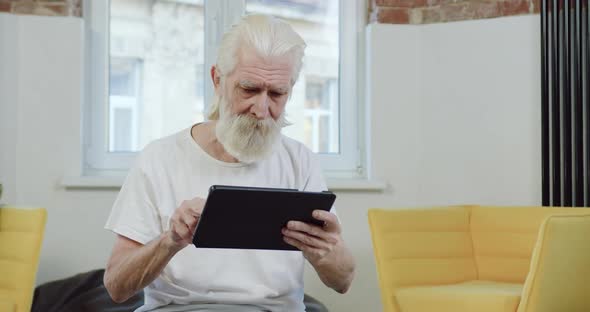 Mature Granddad in white T-shirt Using Tablet Device while Spending Leisure at Home