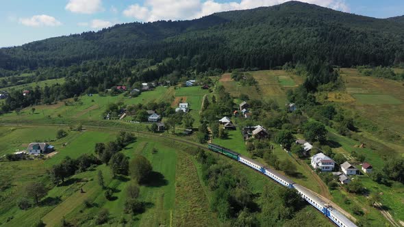 Train Moving on Railroad Tracks in Carpathian Mountains Through the Village Aerial View