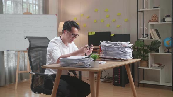 Asian Man Enjoys Playing Game On Smartphone After Working With Documents At The Office