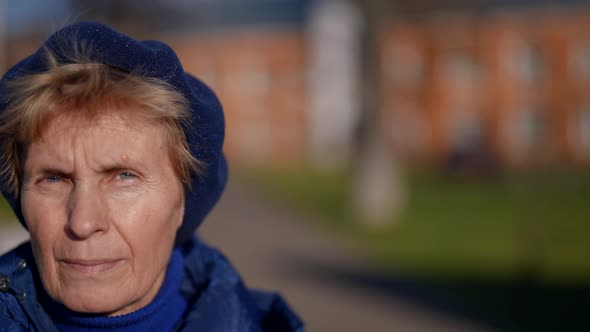 Portrait of an Elderly Woman in a Beret and Blue Clothes Against the Background of Blurred Houses on