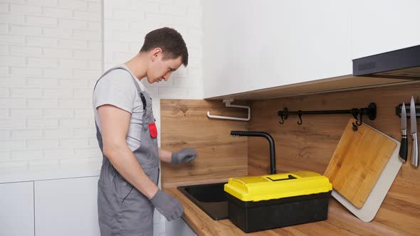 Worker in Jumpsuit with Toolbox Looks for Leakage on Faucet