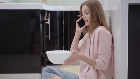 Worried Young Slim Woman Talking on the Phone Sitting on Kitchen Floor As Water Leaking From Ceiling