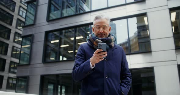 A Grayhaired Man Uses a Mobile Phone Standing in Business Center of the City