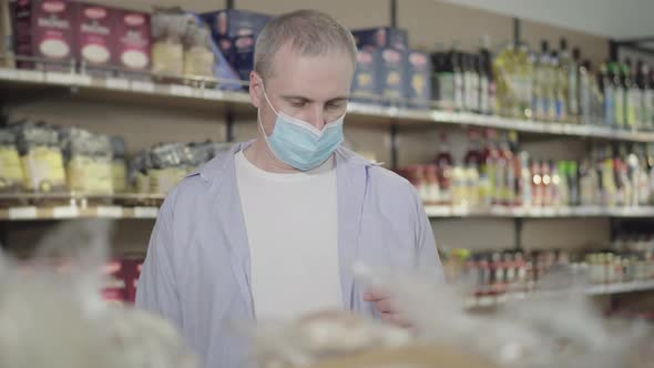 Portrait of Caucasian Man in Face Mask Choosing Products in Grocery. Adult Concentrated Male Buyer