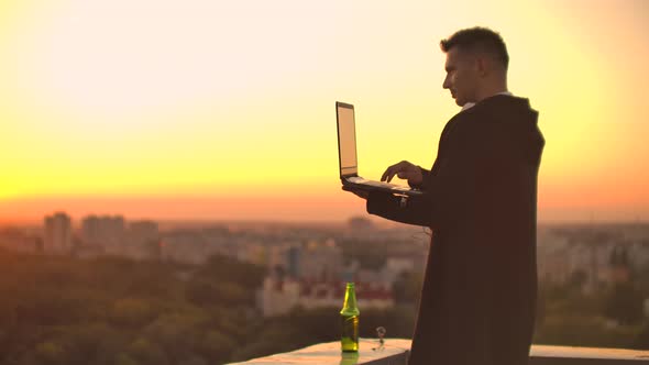 Standing at Sunset on the Roof with a Laptop and a Beer. A Man in a Hoodie Works Having Fun and