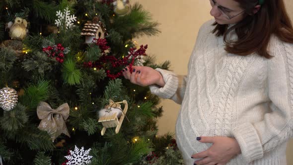 A Young Pregnant Woman in Glasses and a Sweater with Horns on Her Head Examines the Toys on the