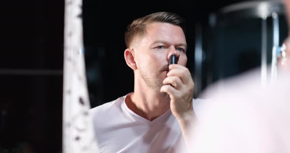 Handsome Young Man Removes Hair From Nose with Trimmer in Front of Mirror