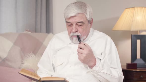 Portrait of Grey-haired Caucasian Man Smoking Pipe and Reading Book at Home. Mature Serious Man