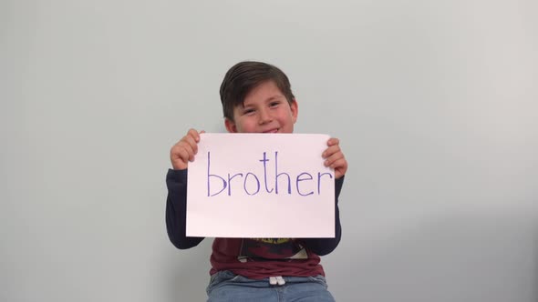 A Boy Shows a Sign with the Word BROTHER on a Grayblue Background A Beautiful Child Laughs and Shows