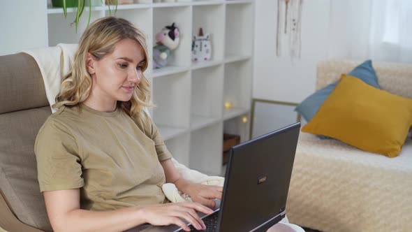 Cute young blonde woman freelancer uses laptop while sitting at home in armchair