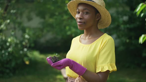 Side View Smiling African American Female Gardener Holding Green Seedling in Pink Gloves Turning