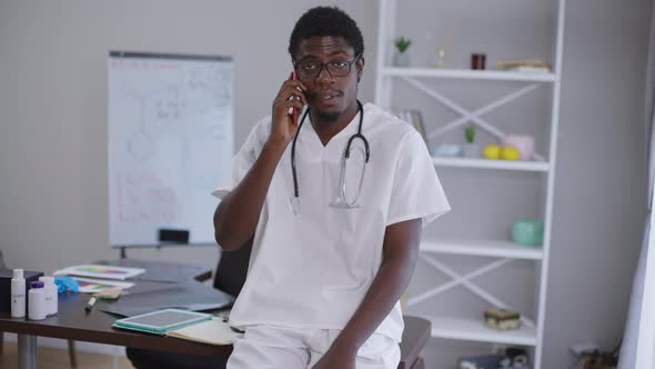 Smart African American Man in Eyeglasses and Doctor Gown Talking on Phone Standing at Table Indoors