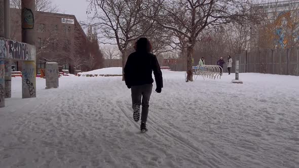 Man wearing parka and boots running on snow covered ground toward park between buildings in Montreal