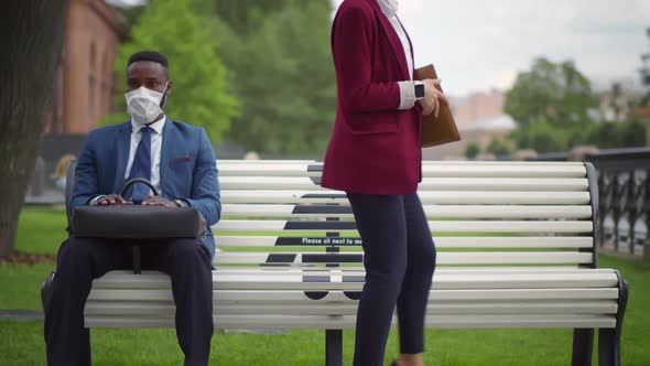 Young Businesspeople in Medical Mask Sitting on Bench with Social Distancing Markup in Public Park.