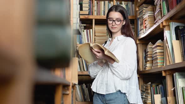 Young Attractive Student in Glasses Turns Pages in the Book Which She Took From Books on Shelves in