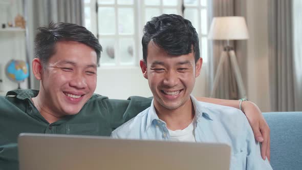 Asian Gentle Gay Couple Using Laptop Computer While Sitting On A Couch, Watching Funny Videos