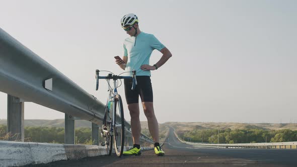 A Guy in Tight Shorts, a Helmet, and Sunglasses Is Standing on the Side of the Road Near the Bike