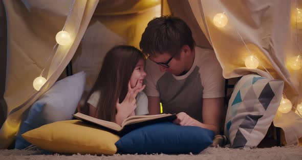 Happy Father and Daughter Lying on Floor in Teepee and Reading Book Together at Home