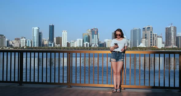 Girl on the Background of the Big City and the Bay Holding a Smartphone in Sunglasses