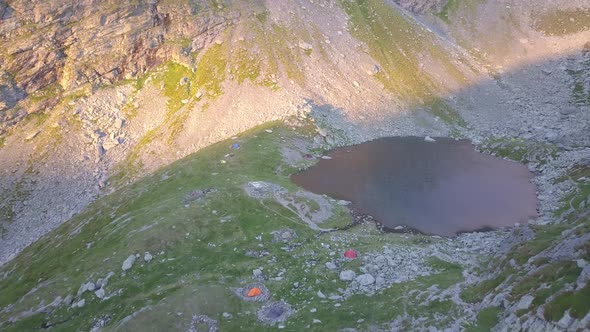 Bright tents of campers beside mountain lake at sunset. Drone flying high over remote high altitude