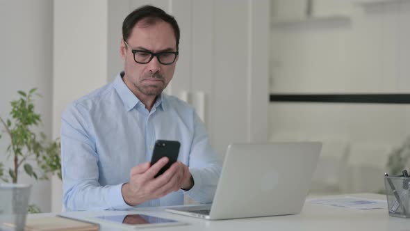 Middle Aged Man Using Smartphone While Working on Laptop