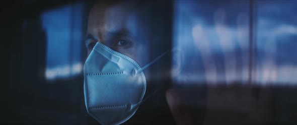 Man with face mask standing with his hand on a window looking out with eyes of hope