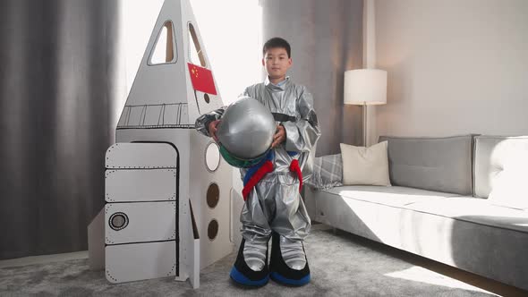 An Asian Boy in an Astronaut Costume Stands Near a Cardboard Model of a Spaceship and Puts on Helmet