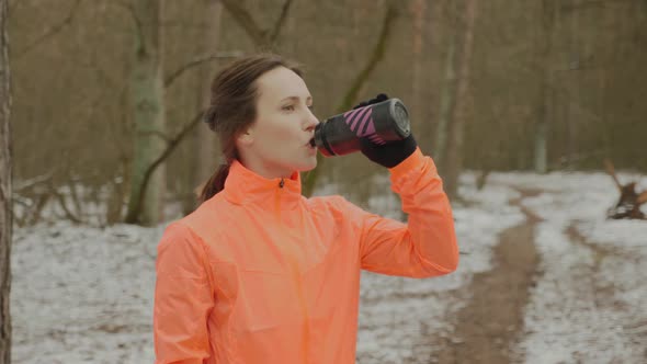 Woman drinking water after running training in winter park wearing orange jacket. Running concept