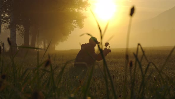 Crouching Man With Camera In Sunlit Meadow At Dawn