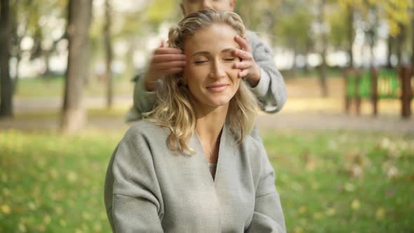 Closeup of Relaxed Beautiful Slim Woman Sitting in Autumn Park As Joyful Boy Running From Background