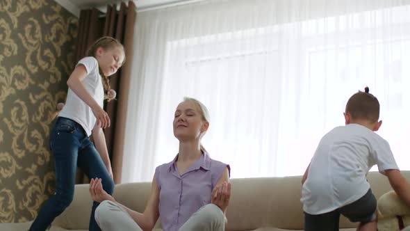 Motherhood, Calm Mother Meditate Sitting on the Couch While the Kids Play and Jump on the Sofa