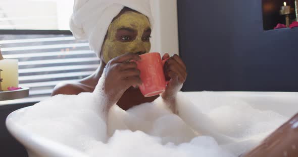 Smiling african american woman with towel and mask taking bath and drinking coffee in bathroom