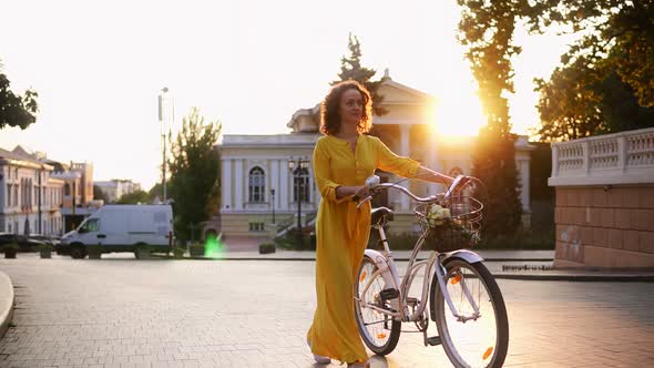 Smiling Brunette Woman in a Long Yellow Dress Walking During the Dawn Holding Her City Bicycle's