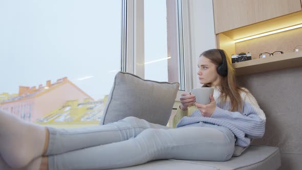 Young woman in headphones sitting at home by the window drinking a hot drink from a mug