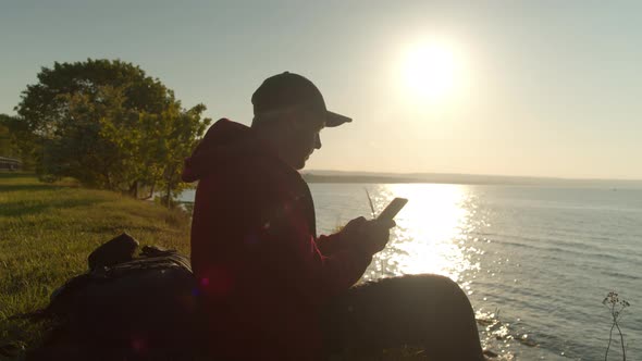 Man Traveler is Sitting on the Ocean or Sea Shore Using a Smartphone