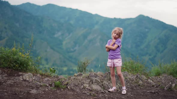 Funny Little Child Touches Cheek with Finger on Rocky Hill