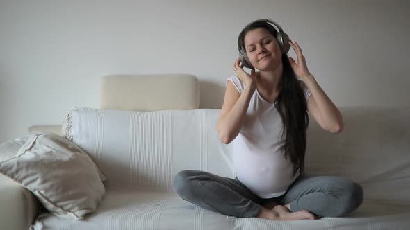 Pregnant Woman Dancing on White Sofa and Listening Music in Headphones at Home. Slow Motion Video