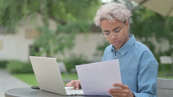 Young African Woman with Laptop Reading Documents in Outdoor Cafe