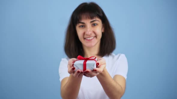 Smiling Brunette Woman Holding Gift Box and Gives It By Hands to Camera on Blue Wall Background