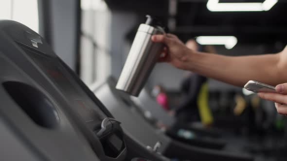 White Man in the Gym Approaches the Treadmill with Bottle and Smartphone in His Hand