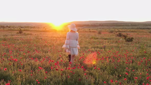 A Young Woman in a White Dress and Hat Walks Towards the Bright Rays of the Sunset Across a Field of