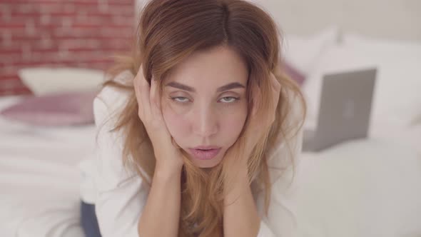 Portrait of an Exhausted Caucasian Girl Sitting at the Bed and Holding Her Head with Hands