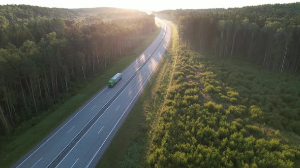 Heavy semi-trailer truck drives through beautiful terrain through the summer woods at dawn.