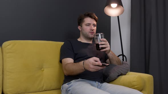 Young Unshaven Guy Watching Movie on TV on Sofa with Drink and Remote Control