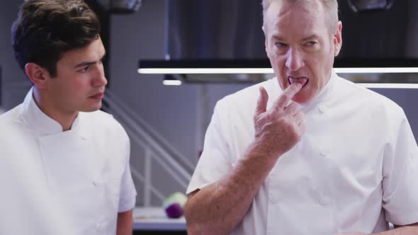 Professional Caucasian male chef in a restaurant kitchen tasting food with a trainee chef