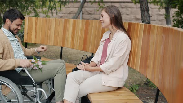 Cheerful Man in Wheelchair Meeting Girlfriend Outdoors in Park Bringing Flowers Talking Enjoying