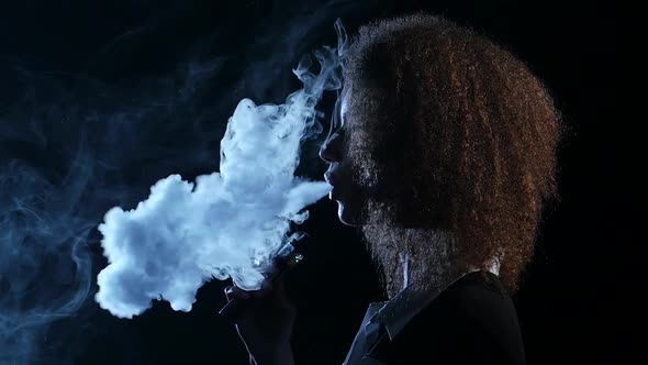 Girl of African American Appearance Smokes an Electronic Cigarette. Black Background. Side View