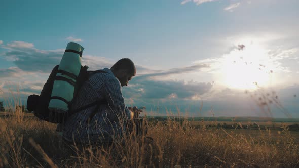 A Young Tourist Man with a Backpack Uses a Mobile Phone. In the Rays of the Setting Sun. Travel