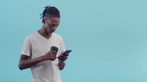 Black man in T-shirt and dreadlocks uses phone and holding a paper cup in hand drinking coffee on a