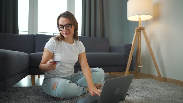 Woman with Glasses is Sitting on the Floor and Makes an Online Purchase Using a Credit Card and